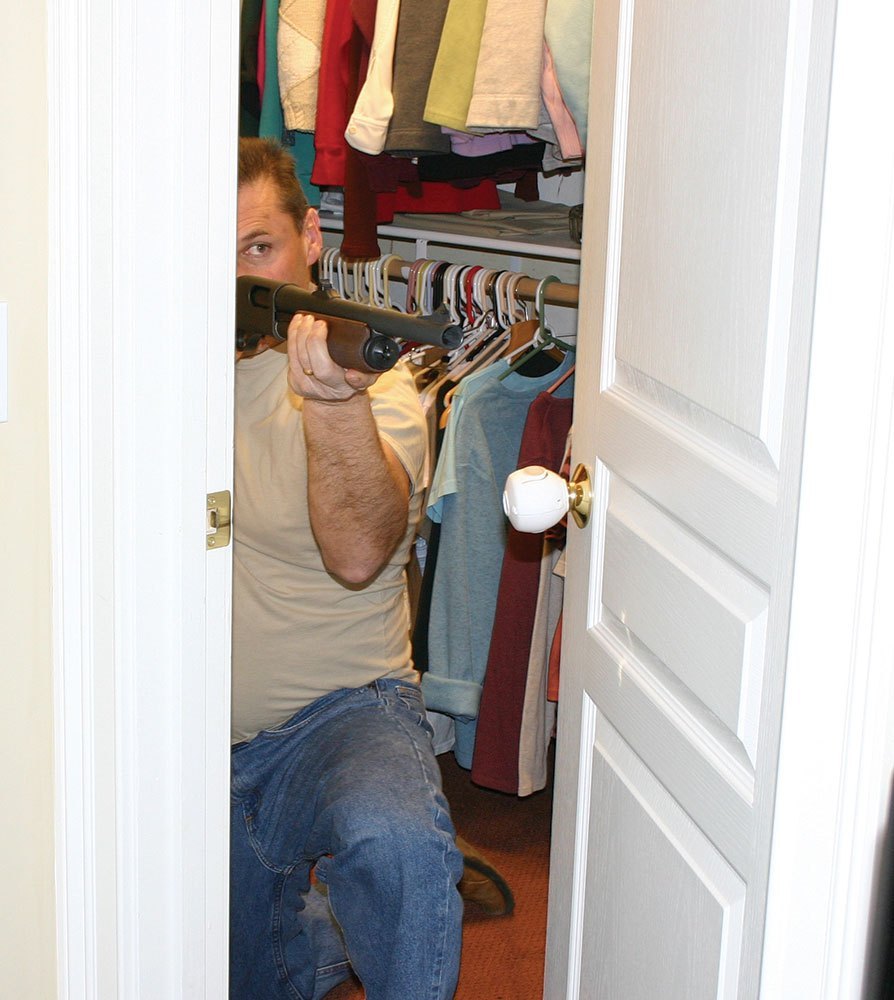 Man kneeling in a closet door aiming a shotgun.